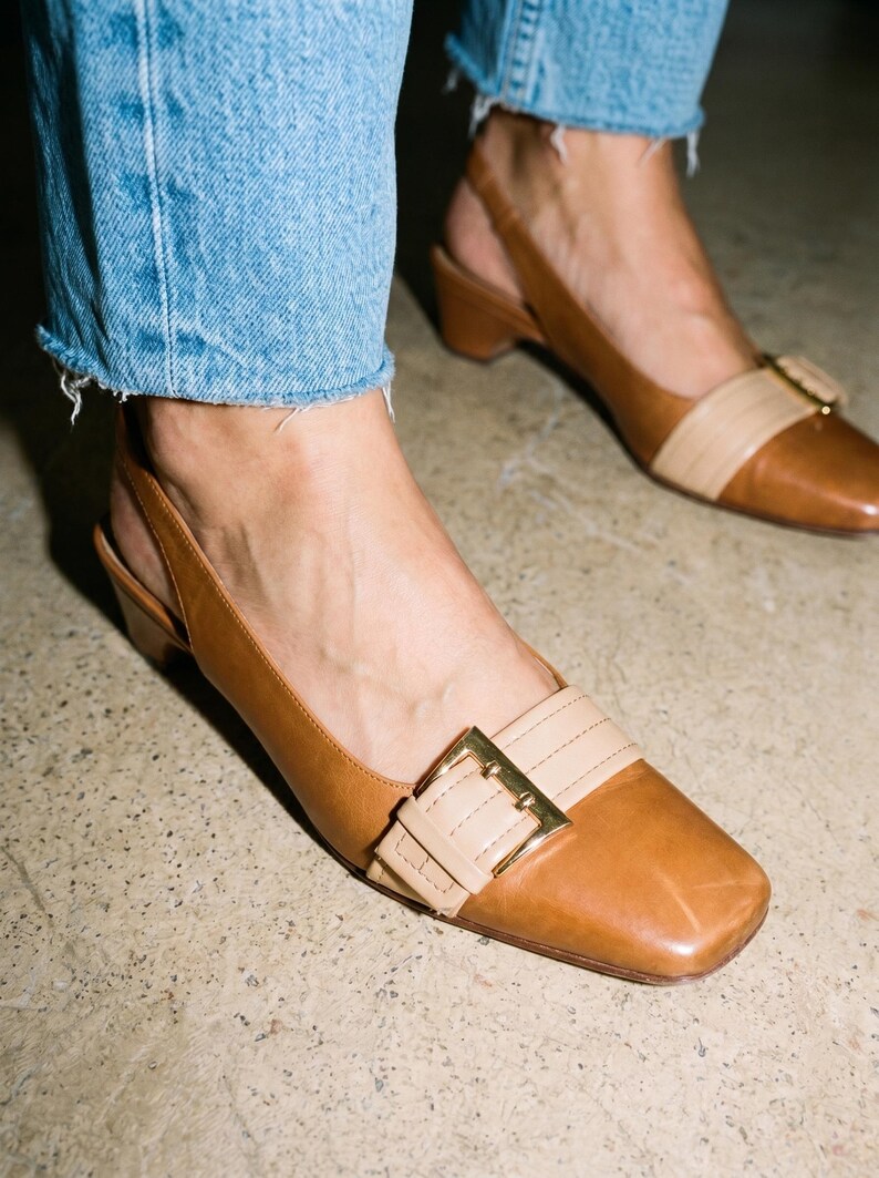K&ouml;nnte beinhalten: Braune Leder-Slingback-Heels mit eckiger Zehenpartie und goldener Schnalle. Die Schuhe haben einen kontrastierenden beigefarbenen Riemen. Die Abs&auml;tze sind niedrig und blockartig. Getragen mit blauen Jeans.