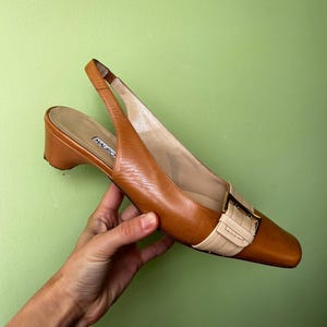 K&ouml;nnte beinhalten: Braune Leder-Slingback-Heels mit eckiger Spitze und kleinem Absatz. Der Schuh hat einen beigefarbenen Riemen mit goldener Schnalle. Der Markenname "MAURO" ist auf der Innensohle sichtbar. Klassisches Design.