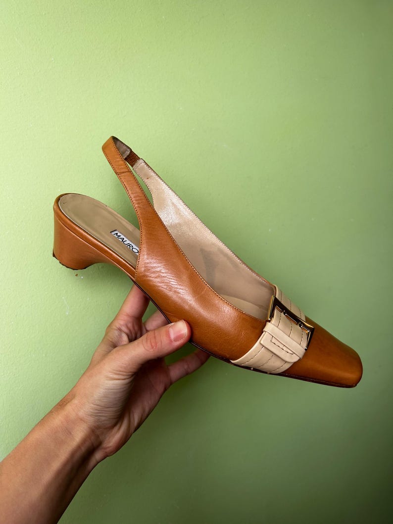 K&ouml;nnte beinhalten: Braune Leder-Slingback-Heels mit eckiger Spitze und kleinem Absatz. Der Schuh hat einen beigefarbenen Riemen mit goldener Schnalle. Der Markenname "MAURO" ist auf der Innensohle sichtbar. Klassisches Design.