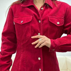 Puede incluir: Una camisa de pana roja con botones, mangas largas y dos bolsillos de solapa. La camisa tiene una silueta ajustada y un cuello puntiagudo.