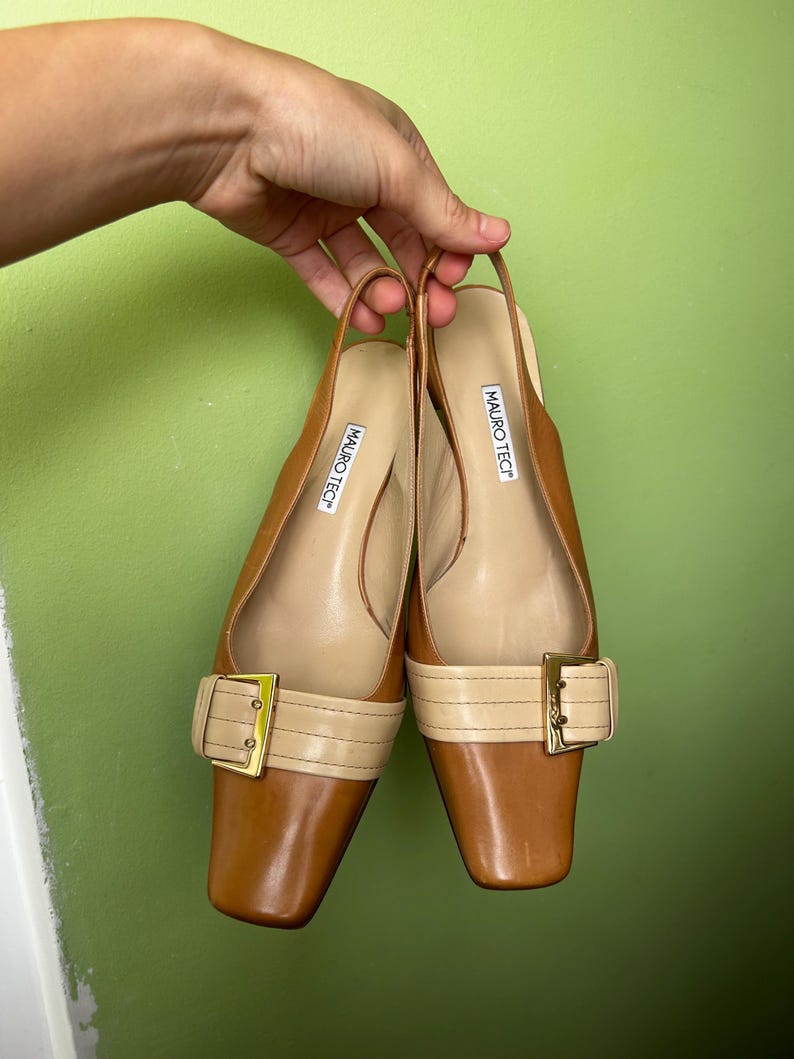 K&ouml;nnte beinhalten: Ein Paar braune Slingback-Schuhe aus Leder mit quadratischen Zehen und einem beigefarbenen Riemen &uuml;ber dem Oberteil, der mit einer goldfarbenen Schnalle befestigt ist. Die Schuhe werden von einer Hand vor einem gr&uuml;nen Hintergrund gehalten. Der Markenname "MAURO TECT" ist auf der Innensohle aufgedruckt.