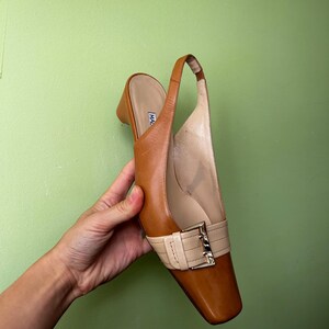 K&ouml;nnte beinhalten: Ein brauner Slingback-Schuh aus Leder mit eckiger Zehenpartie und kleinem Absatz. Der Schuh hat einen cremefarbenen Riemen &uuml;ber dem Oberteil, der mit einer goldfarbenen Schnalle befestigt ist. Der Schuh wird vor einem gr&uuml;nen Hintergrund gehalten.