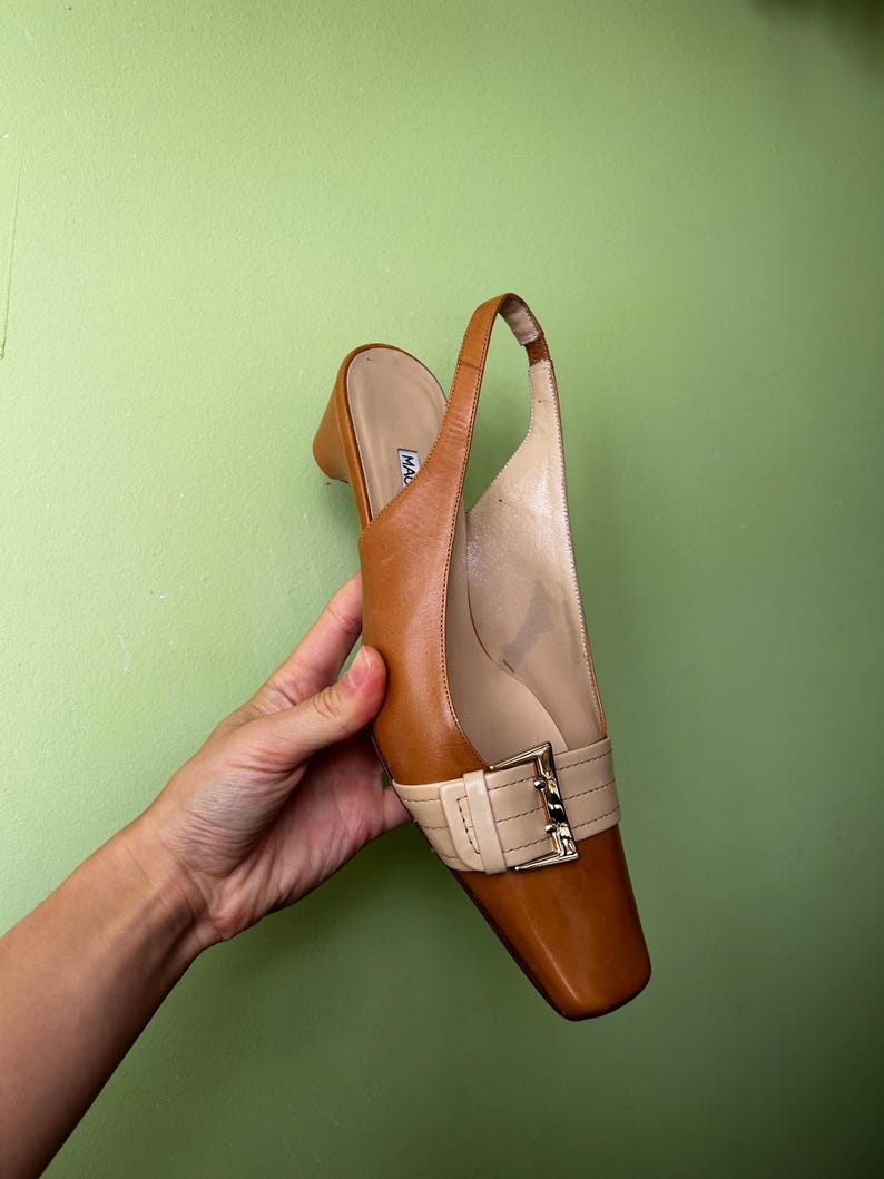 K&ouml;nnte beinhalten: Ein brauner Slingback-Schuh aus Leder mit eckiger Zehenpartie und kleinem Absatz. Der Schuh hat einen cremefarbenen Riemen &uuml;ber dem Oberteil, der mit einer goldfarbenen Schnalle befestigt ist. Der Schuh wird vor einem gr&uuml;nen Hintergrund gehalten.
