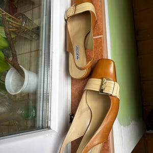 K&ouml;nnte beinhalten: Ein Paar braune und beige Slingback-Heels aus Leder. Die Abs&auml;tze haben eine eckige Spitze, einen beigen Riemen &uuml;ber der Oberseite mit einer goldenen Schnalle und einen Slingback-Riemen. Der Markenname "MAURO TEC" ist auf der Innensohle sichtbar.