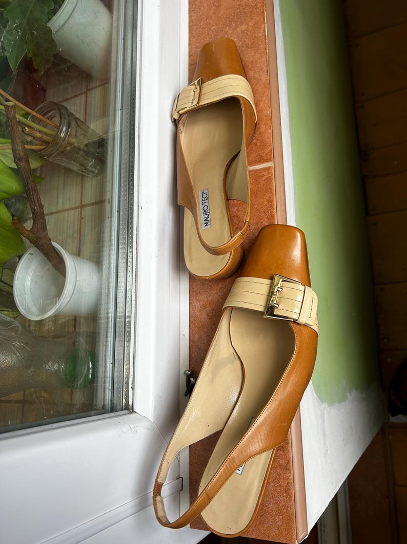 K&ouml;nnte beinhalten: Ein Paar braune und beige Slingback-Heels aus Leder. Die Abs&auml;tze haben eine eckige Spitze, einen beigen Riemen &uuml;ber der Oberseite mit einer goldenen Schnalle und einen Slingback-Riemen. Der Markenname "MAURO TEC" ist auf der Innensohle sichtbar.