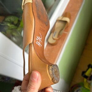 K&ouml;nnte beinhalten: Ein hellbrauner Lederschuh mit spitzer Zehenpartie und kleinem Absatz. Der Schuh hat einen Riemen und eine Schnalle. Die braune Sohle weist Gebrauchsspuren auf. Ein weiterer Schuh ist im Hintergrund zu sehen.