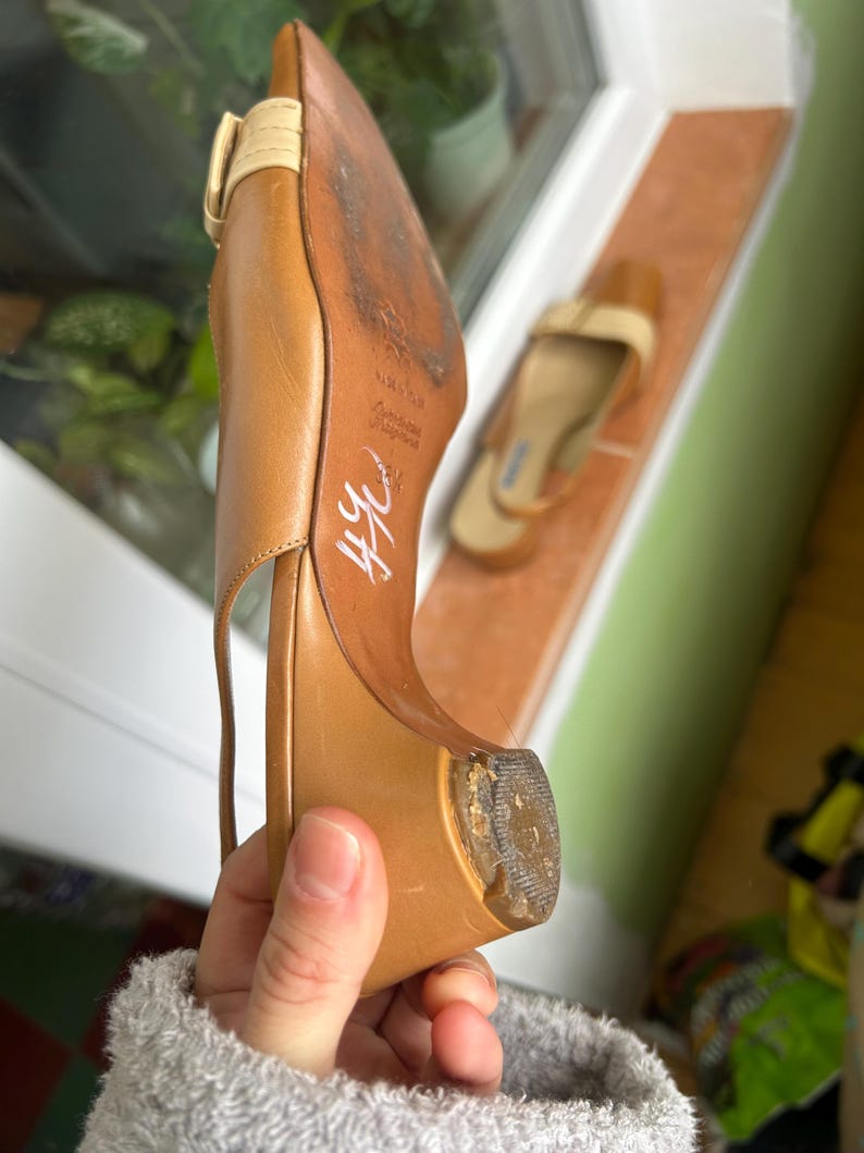 K&ouml;nnte beinhalten: Ein hellbrauner Lederschuh mit spitzer Zehenpartie und kleinem Absatz. Der Schuh hat einen Riemen und eine Schnalle. Die braune Sohle weist Gebrauchsspuren auf. Ein weiterer Schuh ist im Hintergrund zu sehen.