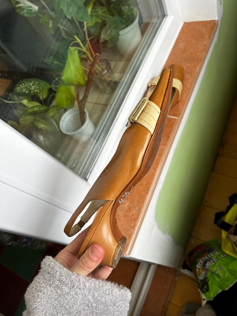 K&ouml;nnte beinhalten: Braune Slingback-Heels aus Leder mit beigem Riemen und Schnalle. Die Schuhe haben einen kleinen Absatz und eine spitze Zehenpartie. Die Schuhe liegen auf einer Fensterbank. Absatzh&ouml;he ca. 5 cm.