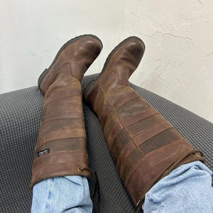 May include: A pair of knee-high brown leather boots featuring horizontal paneling, a rounded toe, and a small black tag on the side with the text K-DRY WATERPROOF. The boots have a lace-up detail at the top and are worn over light blue denim jeans.
