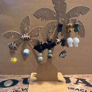 May include: A display of various earrings, including fish, bat, and abstract shapes, hanging from a palm tree-shaped stand. The earrings feature gold-colored hooks and are made of ceramic and other materials. The stand is made of cardboard.