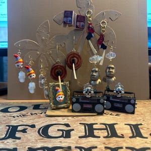 May include: A display of handmade earrings with various designs, including clown faces, miniature books, target shapes, and miniature boomboxes with skull accents. The earrings are arranged on a palm tree-shaped stand.