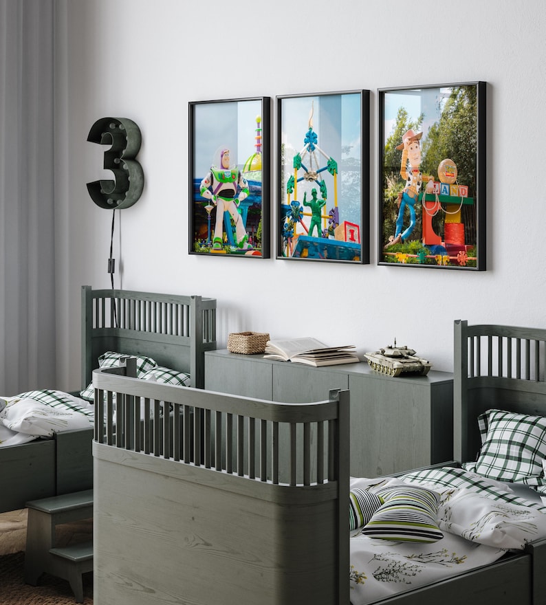 Puede incluir: Tres imágenes enmarcadas de personajes de Toy Story en una habitación infantil. Las imágenes están colgadas en una pared blanca sobre dos camas individuales con cabeceros de madera verde. Las camas están hechas con ropa de cama blanca y verde. Un tanque de juguete está en un estante sobre las camas.
