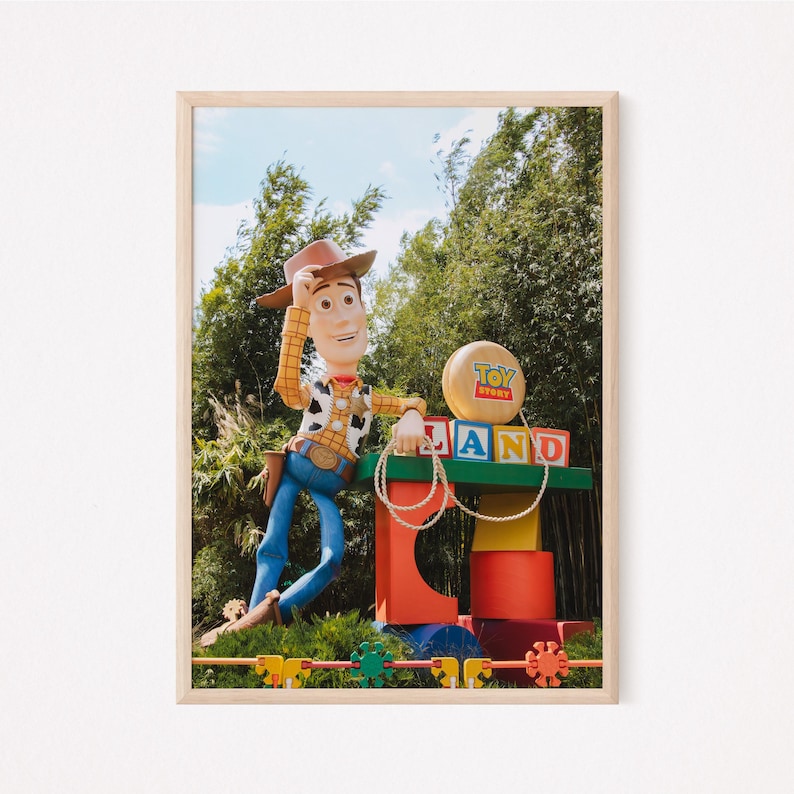 Puede incluir: Un gran Woody de Toy Story de dibujos animados se encuentra junto a un cartel colorido que dice "Toy Story Land". El cartel est&aacute; hecho de grandes bloques coloridos. Woody lleva su caracter&iacute;stico sombrero de vaquero y chaleco. Se apoya en el cartel con la mano derecha en la cadera.