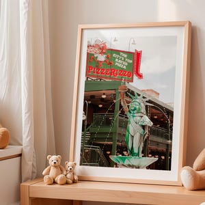 Puede incluir: Una fotografía enmarcada de una estatua verde de la Estatua de la Libertad sosteniendo una rebanada de pizza frente a un edificio con un letrero que dice "The City's Top Rated Pizza, Pizzeriazzo".