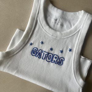 Pode incluir: Top branco canelado com a palavra "GATORS" bordada em azul, juntamente com detalhes de estrelas azuis. O top tem decote redondo e design sem mangas. O tecido tem canelado vertical.