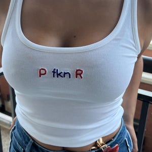 May include: White ribbed vest top with the embroidered text "P fkn R" in red and blue. The top is worn with blue denim jeans. A gold and red floral brooch is attached to the jeans.