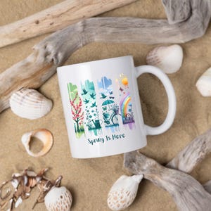 May include: White ceramic mug with a colorful spring-themed design. The design includes a tree, birds, an umbrella, a bicycle, and a rainbow. The text "Spring Is Here" is printed on the mug.