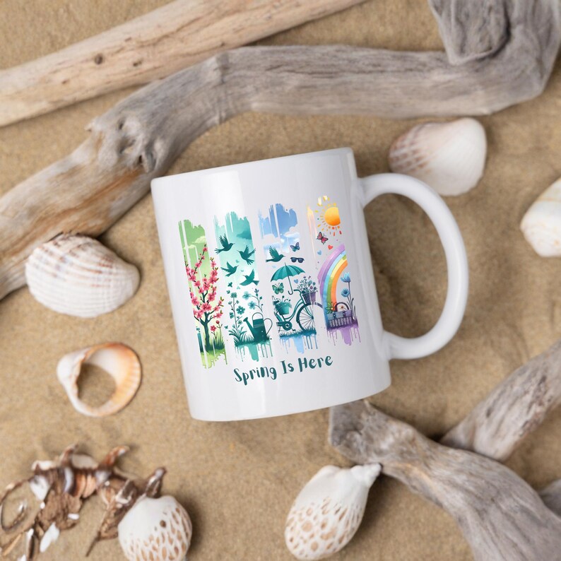 May include: White ceramic mug with a colorful spring-themed design. The design includes a tree, birds, an umbrella, a bicycle, and a rainbow. The text "Spring Is Here" is printed on the mug.