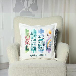 May include: White square pillow with a colorful spring-themed design. The design includes images of a flowering tree, birds, a watering can, a bicycle with a basket of flowers, a rainbow, and a sun. The text "Spring Is Here" is printed on the pillow.