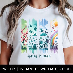 May include: White t-shirt featuring a colorful spring-themed design. The design includes images of a flowering tree, birds, butterflies, a watering can, a bicycle, a rainbow, and the text "Spring Is Here".