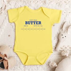 May include: A yellow baby onesie with a blue graphic of a butter label that reads "Sweet Cream Butter, Grade AA, Net Wt. 4 oz. (113g)" and a scale of tablespoons.