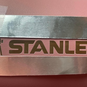 May include: A silver metal plate with a gold logo that says "STANLEY" with a registered trademark symbol. The logo is on a pink background.
