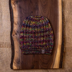 May include: A colorful knitted beanie hat with a brown leather tag that says "hide". The hat is sitting on a wooden cutting board.