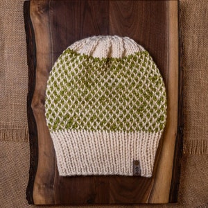 Könnte beinhalten: Eine gestrickte Mütze mit einem grünen und weißen Rautenmuster. Die Mütze liegt auf einer Holzoberfläche.