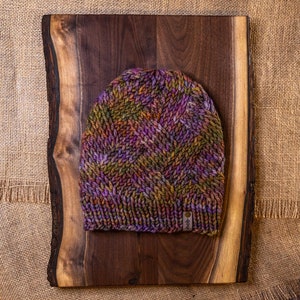 May include: A purple, green, and brown knitted beanie hat with a brown leather tag. The hat is sitting on a wooden surface.