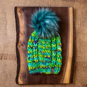 Könnte beinhalten: Eine handgestrickte Mütze in Grün-, Gelb- und Lilatönen, mit einem großen Bommel aus türkisfarbenem Kunstfell. Die Mütze wird auf einem Holzbrett mit Jutegrund präsentiert. Ein kleines Etikett ist auf die Mütze genäht.