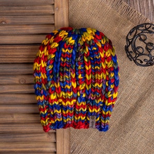 Könnte beinhalten: Eine bunte Strickmütze mit einem roten, gelben, blauen und grünen Farbmuster. Die Mütze ist aus dickem Garn gefertigt und hat eine grob gestrickte Textur.