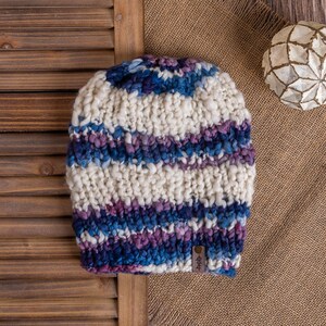 Könnte beinhalten: Eine gestrickte Mütze mit weißen und blauen Streifen und einem braunen Lederanhänger. Die Mütze liegt auf einer braunen Jutestoffe mit einer weißen Dekorkugel.