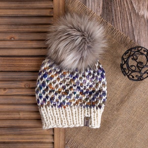 May include: A white and multi-colored knitted beanie with a faux fur pom pom. The beanie is sitting on a brown burlap surface with a wooden shutter in the background.