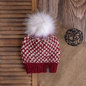 May include: Red and white knitted beanie with a white faux fur pom pom. The beanie is on a brown wooden surface with a burlap fabric.