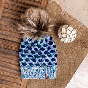 May include: A blue and purple knitted beanie with a brown faux fur pom pom. The beanie has a textured pattern and a small brown leather tag with the word "Help" on it.