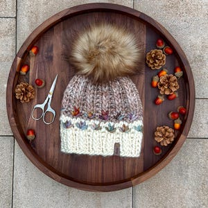 Könnte beinhalten: Eine handgestrickte Wintermütze mit einem Kunstfell-Bommel, präsentiert auf einem Holztablett mit Eicheln, Tannenzapfen und einer kleinen Schere. Die Mütze hat einen cremefarbenen Rand und eine braun-cremefarbene Krone mit bunten Blättern.