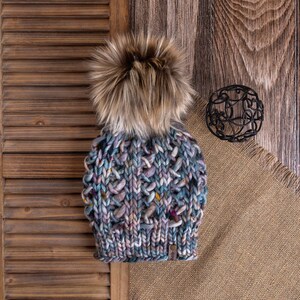 Pode incluir: Um gorro de malha azul, roxo e castanho com um pompom castanho felpudo. O gorro está sobre uma superfície de madeira castanha com um pedaço de tecido de juta.