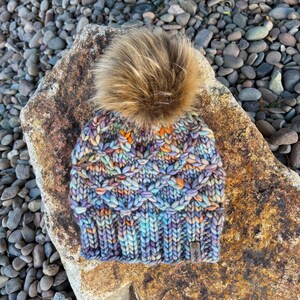 Pode incluir: Um gorro de malha colorido com um pompom marrom felpudo. O gorro tem um padrão de malha de cabo e é feito com uma variedade de tons de azul, roxo, laranja e verde. O gorro tem uma pequena etiqueta de couro com a palavra "#feb" nela.