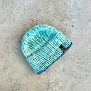 Könnte beinhalten: Eine hellblaue Strickmütze mit blauem und violettem Saum. Die Mütze hat ein kleines braunes Etikett mit dem Wort "shop" darauf.