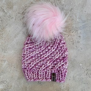 May include: A pink knitted beanie with a fluffy pink pom pom on top. The beanie has a  "Heb-" leather tag attached to the side.