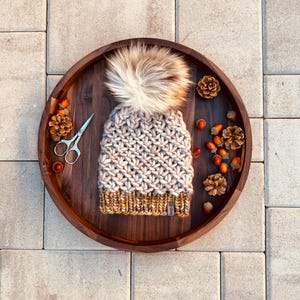 Könnte beinhalten: Eine handgestrickte Mütze mit einem Kunstfell-Bommel, präsentiert auf einem Holztablett mit herbstlichen Dekorationen. Die Mütze hat ein cremefarbenes und beiges Farbschema mit einem senfgelben Rand. Eine Schere und Eicheln befinden sich ebenfalls auf dem Tablett.