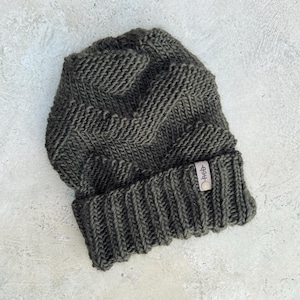 Könnte beinhalten: Eine dunkelgrüne Strickmütze mit einem kleinen Lederlabel, auf dem "Hebb" steht.