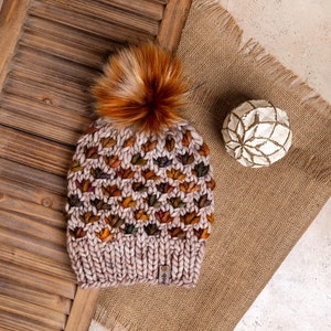 Könnte beinhalten: Eine Strickmütze mit Bommel in Braun-, Orange- und Lilatönen. Die Mütze hat ein strukturiertes Muster und ein kleines Etikett mit der Aufschrift "#fib".