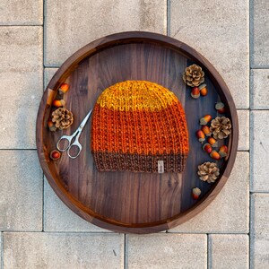 Könnte beinhalten: Handgestrickte Mütze in Herbstfarben Gelb, Orange und Braun. Die Mütze liegt auf einem Holztablett mit Tannenzapfen, Eicheln und einer kleinen Schere. Ein kleines Etikett ist an der Mütze befestigt.