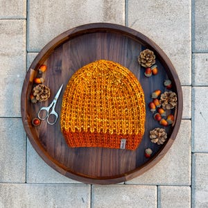 Könnte beinhalten: Eine handgestrickte Mütze in Orange- und Gelbtönen mit Rippenmuster. Die Mütze wird auf einem Holztablett mit Tannenzapfen, Eicheln und einer kleinen Schere präsentiert. Die Mütze hat ein kleines Etikett.