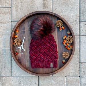 Könnte beinhalten: Burgunderfarbene Strickmütze mit großem Kunstfell-Bommel, präsentiert auf einem Holztablett mit Eicheln, Tannenzapfen und einer kleinen Schere. Die Mütze hat ein strukturiertes Strickmuster und ein kleines Etikett.