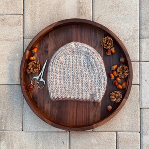 Könnte beinhalten: Eine handgestrickte Mütze in Grau- und Rosatönen liegt auf einem dunklen Holztablett. Das Tablett ist mit Tannenzapfen, Eicheln und einer kleinen Schere dekoriert. Die Mütze hat eine gerippte Struktur und ein kleines Etikett.