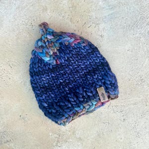 Könnte beinhalten: Eine blaue Strickmütze mit einem mehrfarbigen Muster und einem kleinen braunen Lederanhänger mit der Aufschrift "sheep".