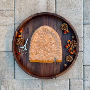 Könnte beinhalten: Eine handgestrickte Mütze in Orange- und Cremetönen mit einem kontrastierenden senfgelben Bund. Die Mütze liegt auf einem dunklen Holztablett, umgeben von Tannenzapfen, Eicheln und einer kleinen Schere. Ein kleines Etikett ist an der Mütze befestigt.