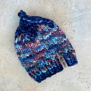 Könnte beinhalten: Eine gestrickte Mütze in Blau, Lila und Rosa mit einem braunen Lederanhänger, auf dem "#bff" steht.